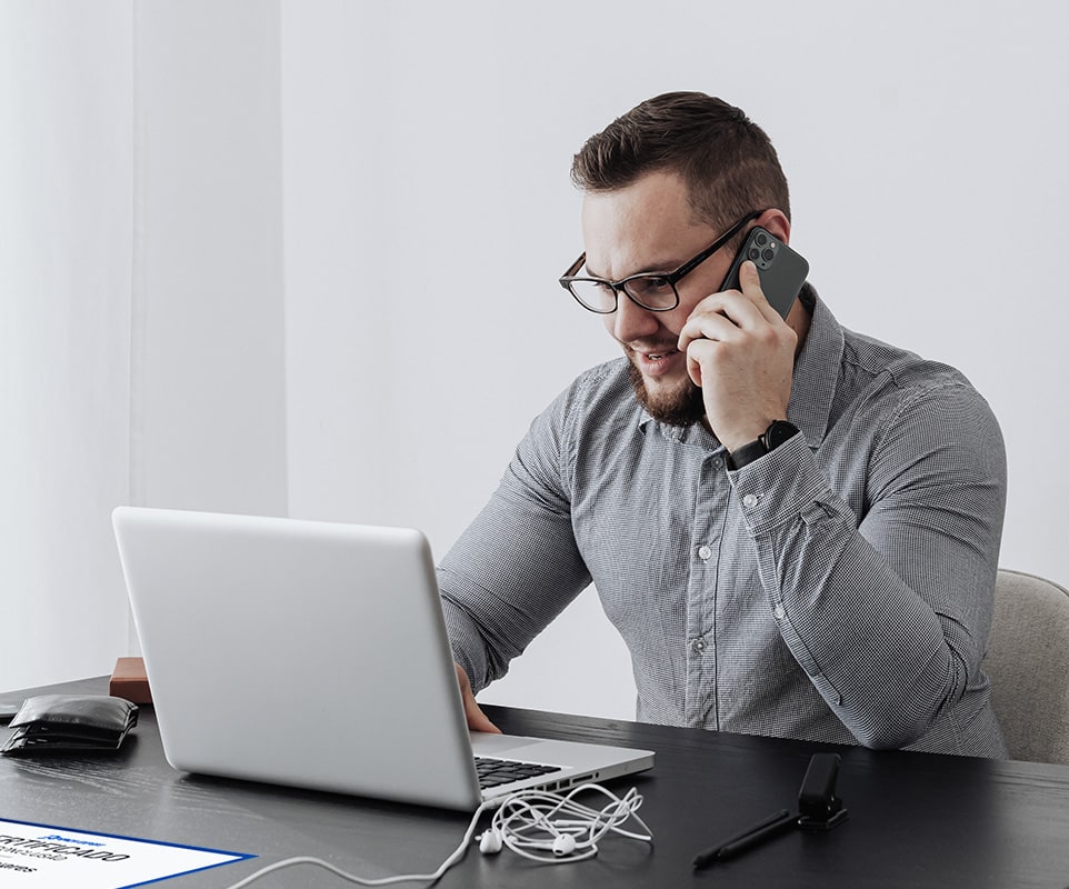 A imagem contém um homem, em um escritório, manuseando o laptop e atendendo o telefone